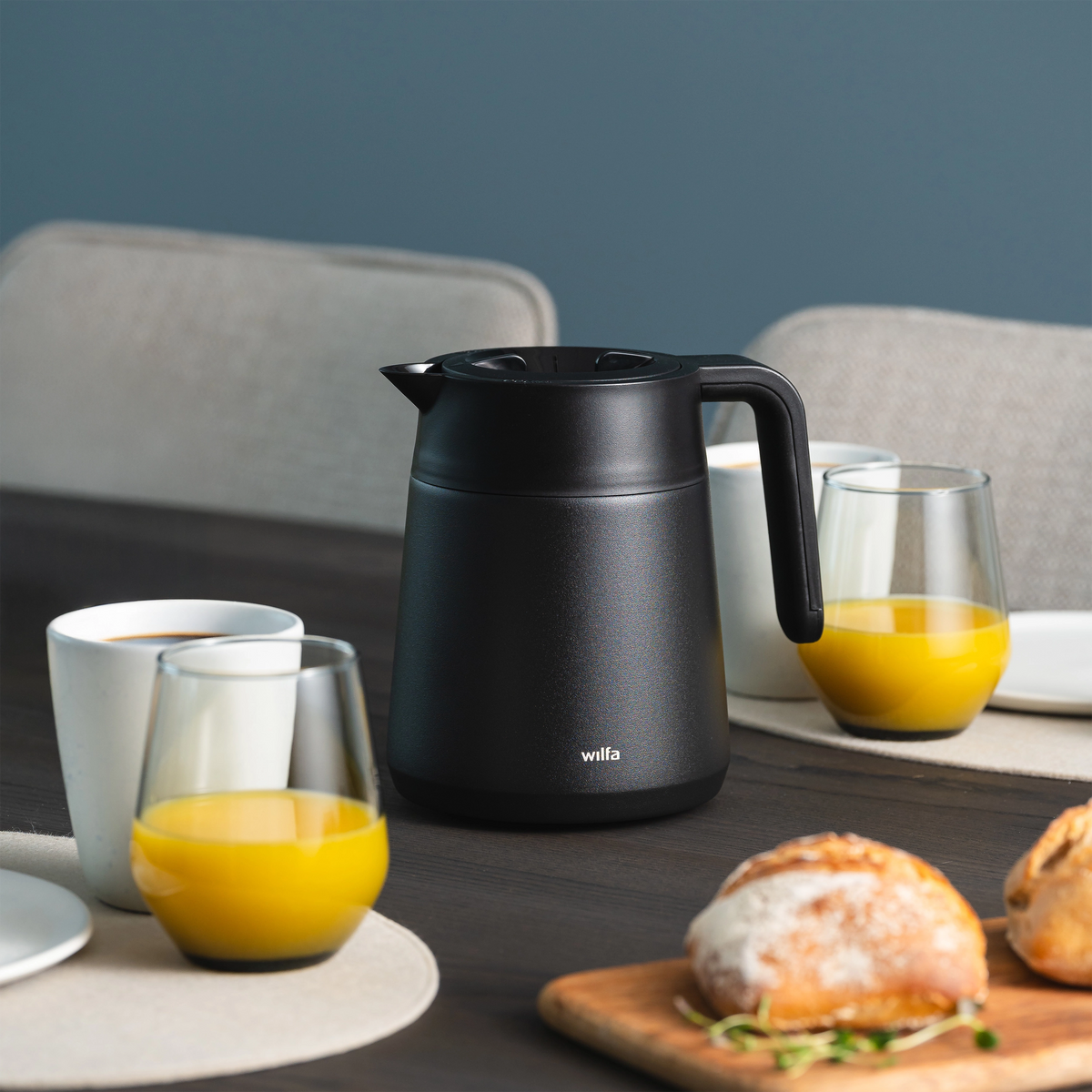 Black Wilfa thermal carafe on a breakfast table with a cup of coffee, glasses of orange juice and fresh bread rolls on a cutting board.