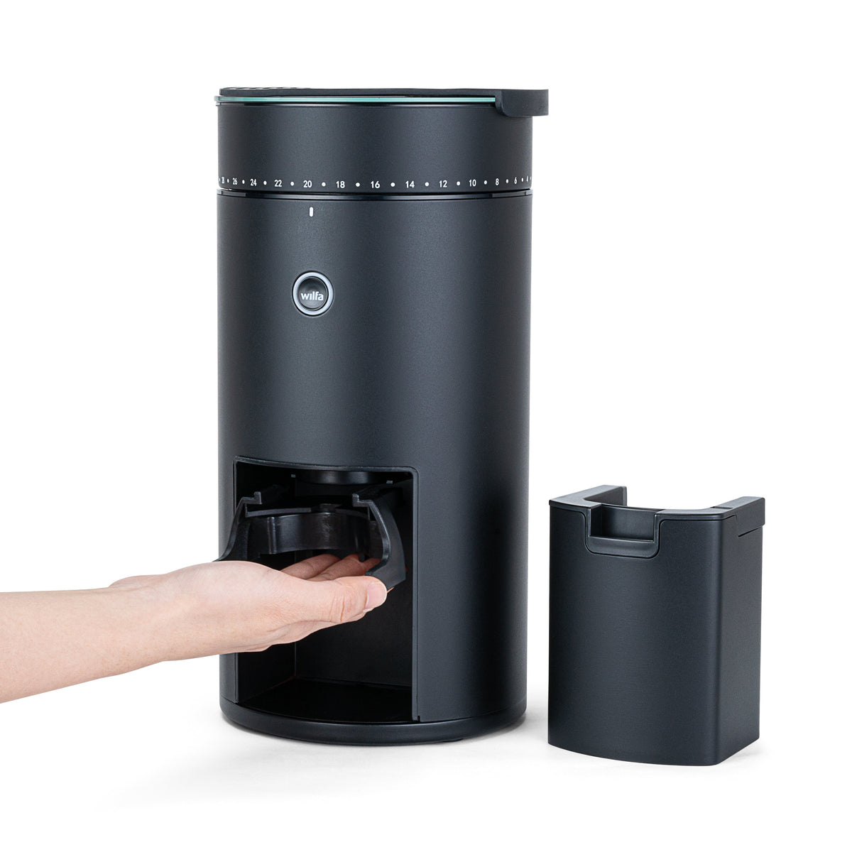 A hand removing the grounds container from the Wilfa Uniform Evo black coffee grinder, with a second container standing beside it.