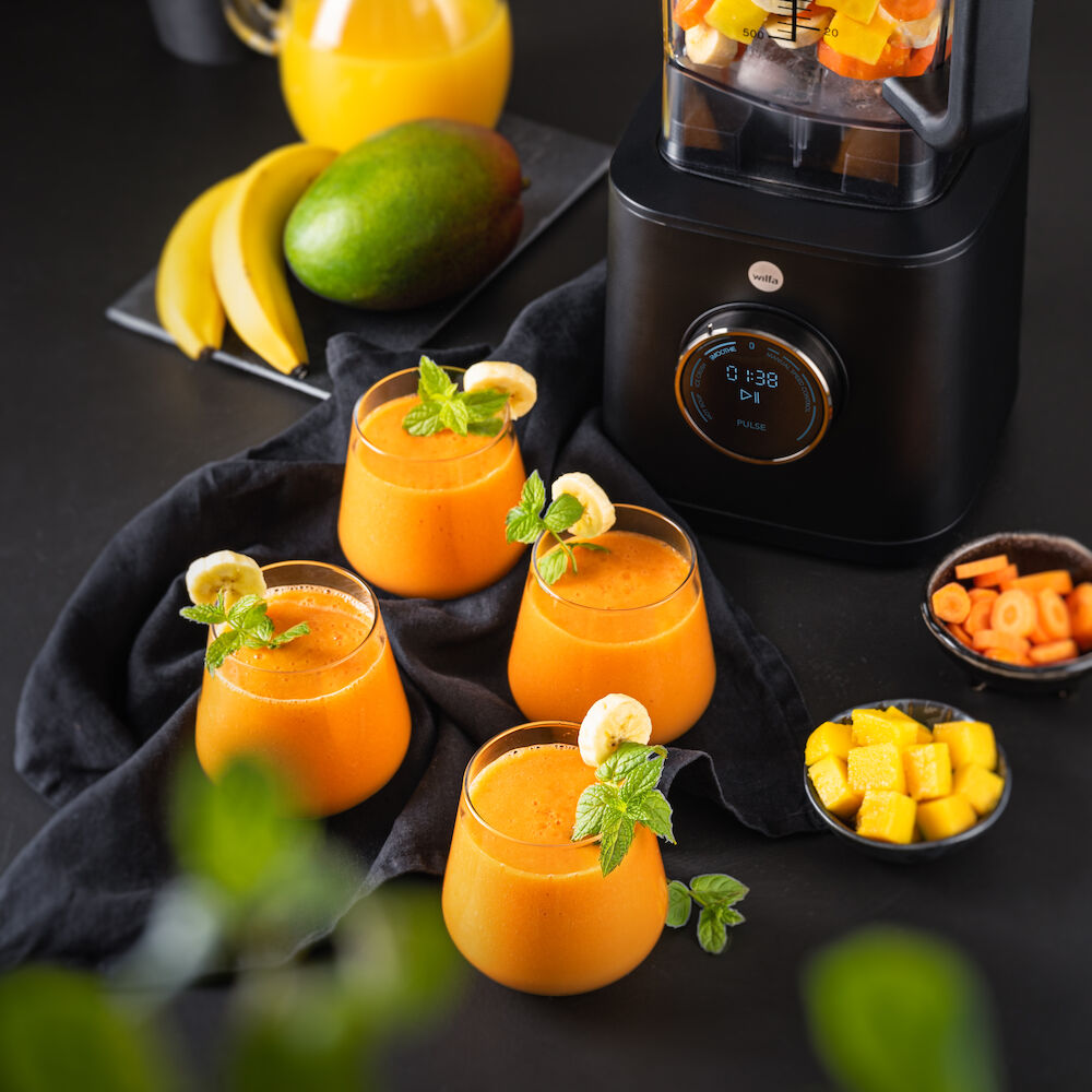 Wilfa Powerfuel XL black blender on a dark table next to three glasses of mango smoothie garnished with mint and banana, with bowls of diced mango and carrot.
