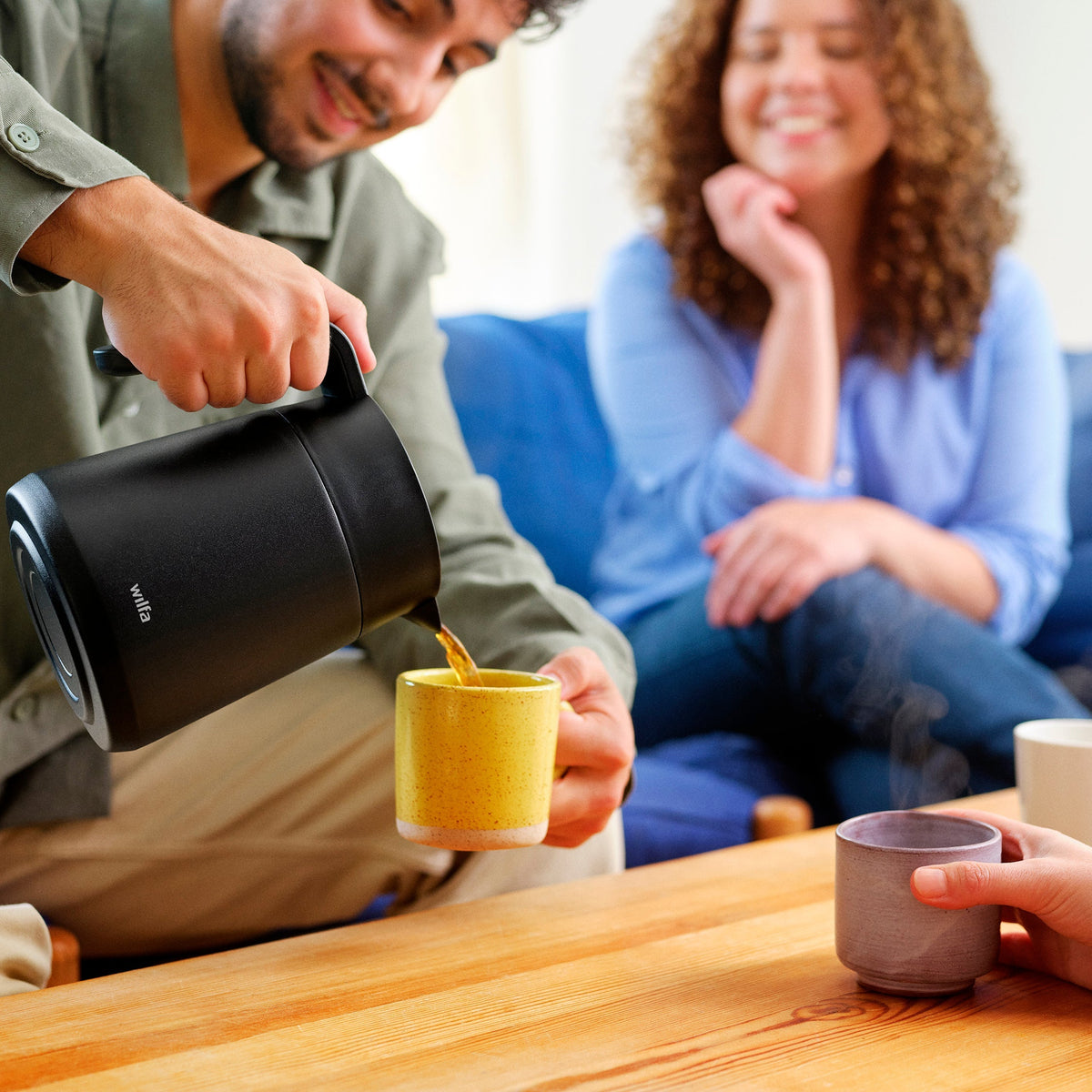 A person pouring coffee from the Wilfa thermal carafe into a yellow mug while two smiling women sit on a couch in the background.