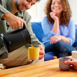 A person pouring coffee from the Wilfa thermal carafe into a yellow mug while two smiling women sit on a couch in the background.