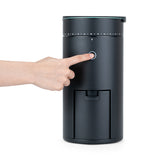 A finger pressing the round start button on the front of the Wilfa Uniform Evo black coffee grinder.