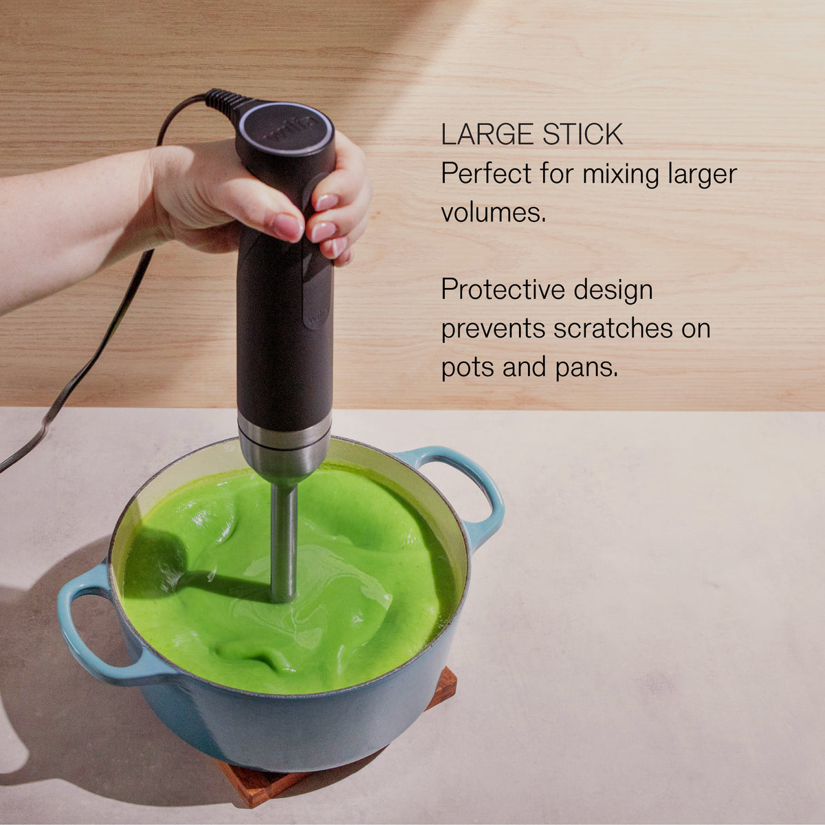 A hand using the Wilfa Prostick immersion blender to blend green soup in a blue pot, with the text 'Large stick: perfect for mixing larger volumes. Protective design prevents scratches on pots and pans.'
